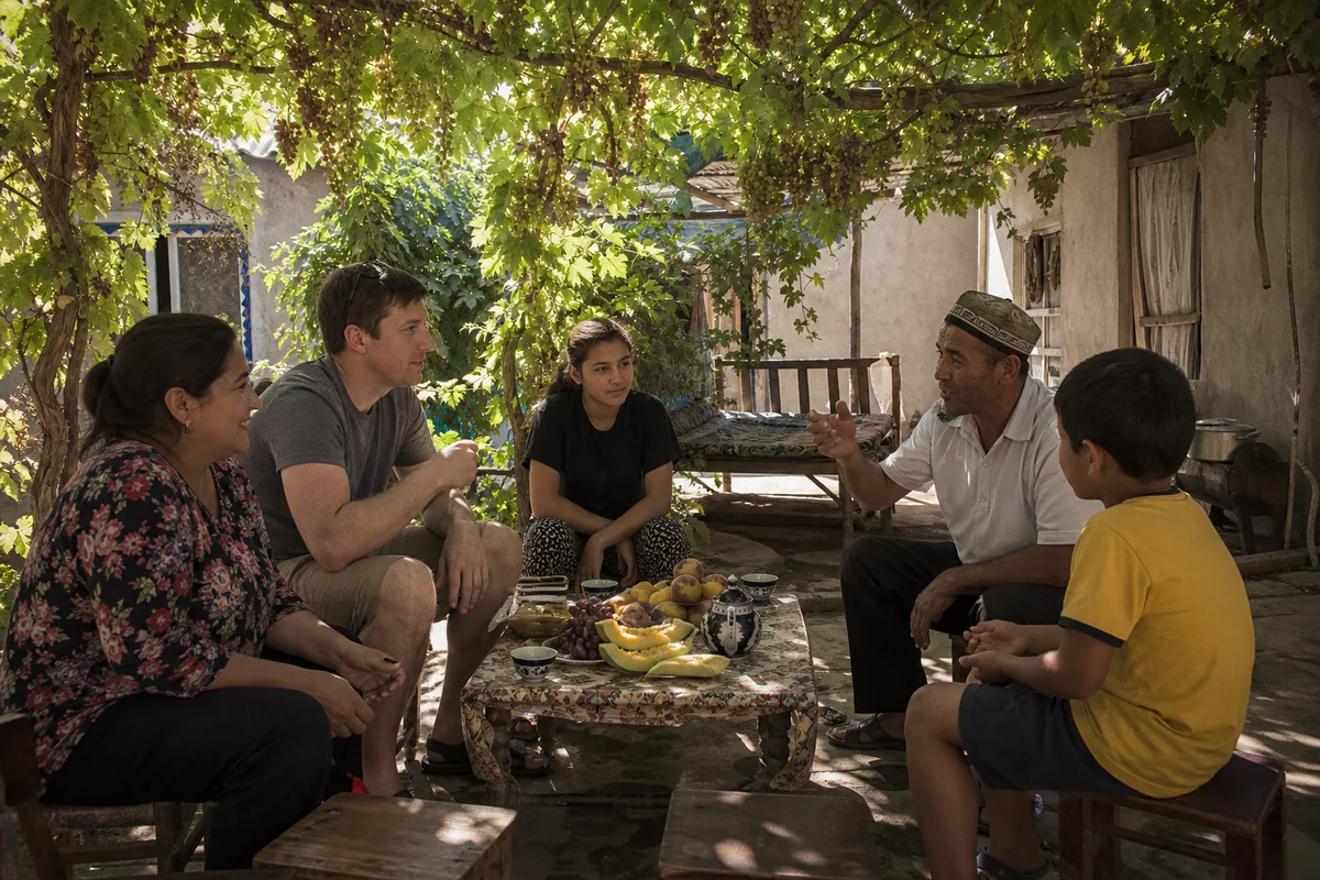 International family sharing tea with a local host under a Turpan grape trellis